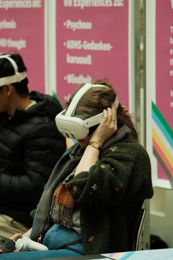 VR-Simulation: Eine sitzende Person trägt eine Virtual-Reality-Brille und hält einen Controller in der Hand, im Hintergrund stehen Informationsplakate.
